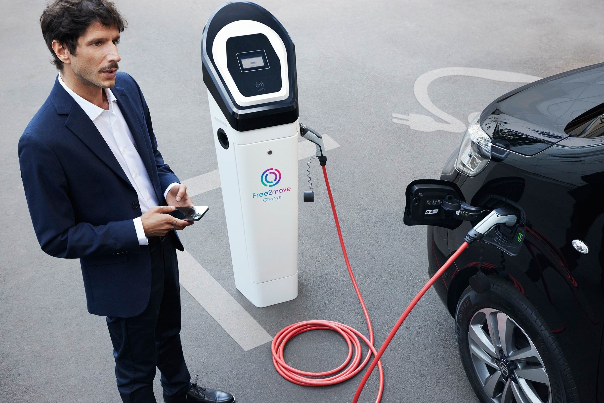 Elektrisch voertuig aan het opladen aan een openbaar laadstation terwijl een smartphone-app wordt gebruikt om het laden te beheren met Free2move Charge